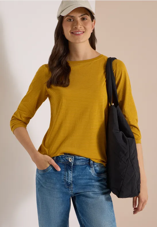 Junge Frau in gelbem Langarmshirt, Jeans, Baseballcap und schwarzer Umhängetasche lächelt.