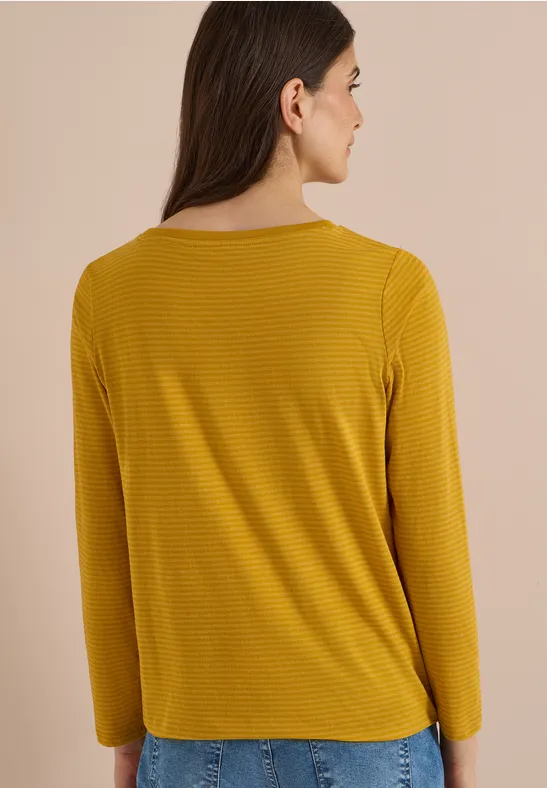 Frau von hinten in senfgelbem, langärmligem Shirt und blauer Jeans vor beigem Hintergrund.