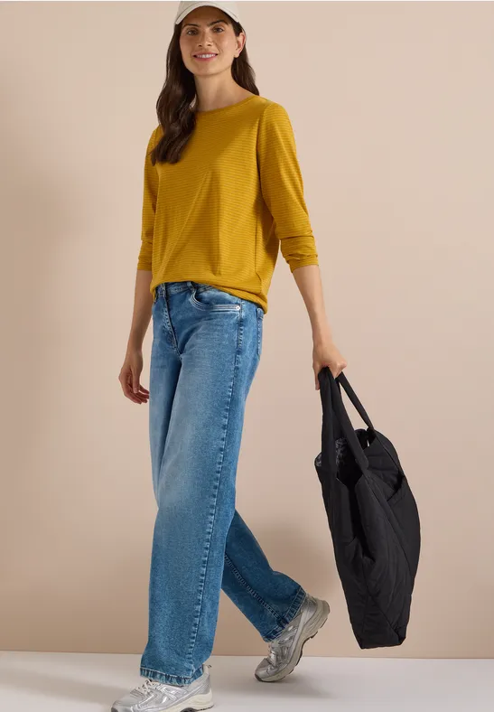 Frau in gelbem 3/4-Shirt, blauen Jeans und Sneakern trägt eine große schwarze Tasche.