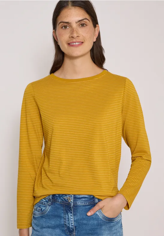 Frau in gelbem, fein gestreiftem Langarmshirt und Jeans, lächelnd vor hellem Hintergrund.