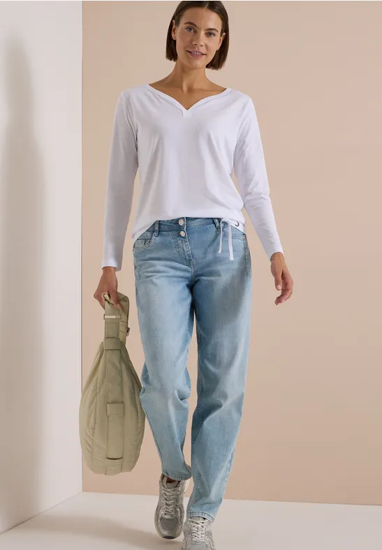 Frau in weißem V-Ausschnitt-Shirt, hellblauen Jeans und silbernen Sneakers hält grüne Tasche vor beigem Hintergrund.