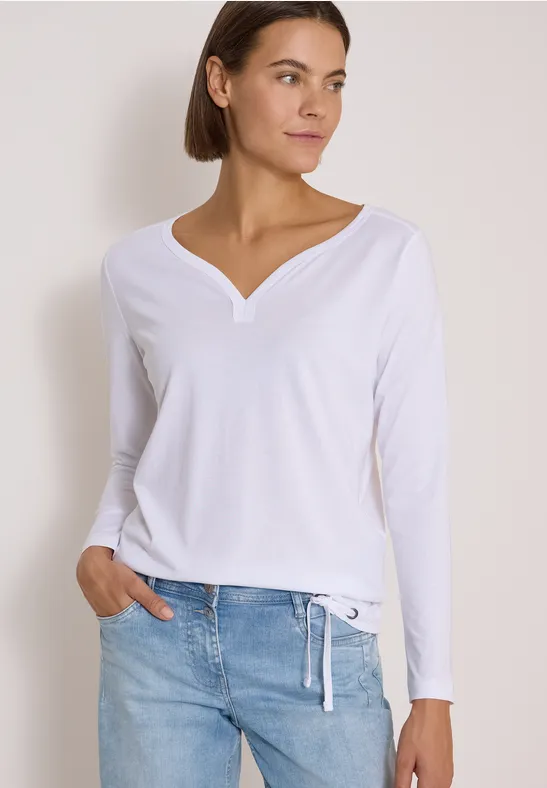 Frau in weißem Langarmshirt mit V-Ausschnitt und Jeans, locker posierend vor heller Wand.