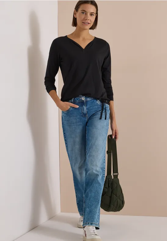 Frau lehnt an Wand, trägt schwarzes V-Shirt, helle Jeans und hält eine grüne Tasche.
