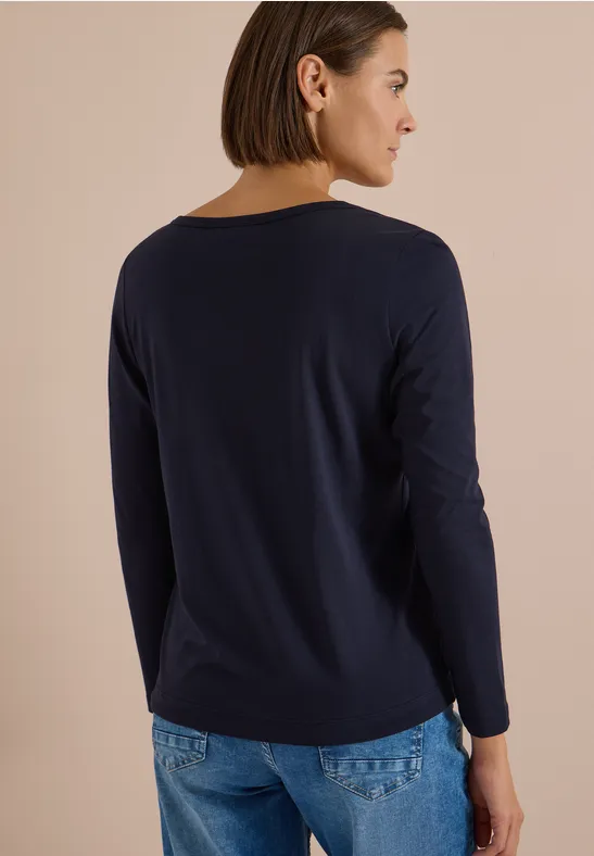 Frau von hinten in dunkelblauem Langarmshirt und Jeans vor neutralem Hintergrund.