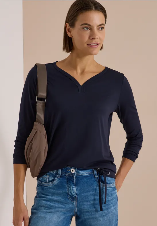 Frau in dunkelblauem 3/4-Ärmel-Shirt mit V-Ausschnitt, Jeans und Schultertasche, lächelnd seitlich blickend.