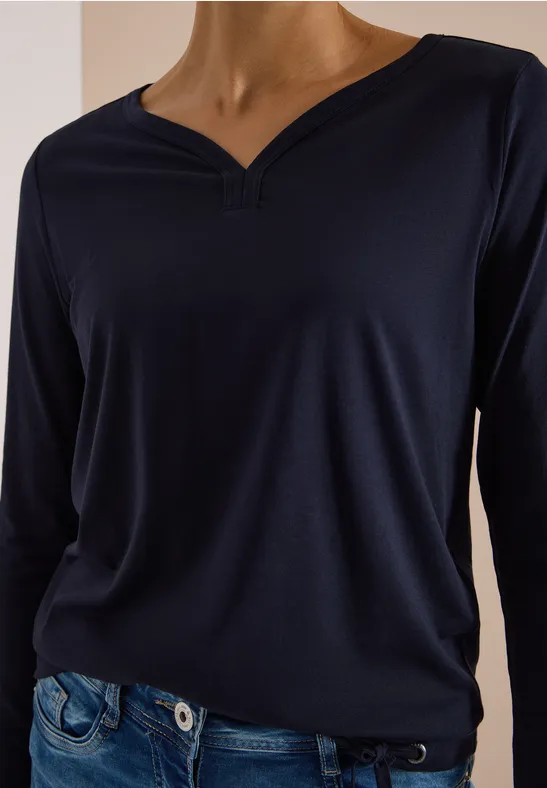 Frau trägt ein dunkelblaues Langarmshirt mit V-Ausschnitt und Jeans vor hellem Hintergrund.