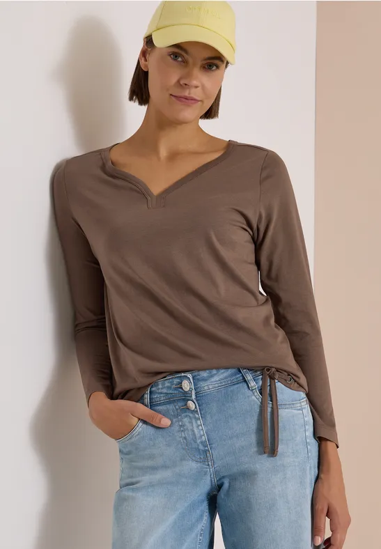 Frau trägt braunes V-Neck-Shirt mit seitlichem Kordelzug, blaue Jeans und gelbe Cap vor neutralem Hintergrund.