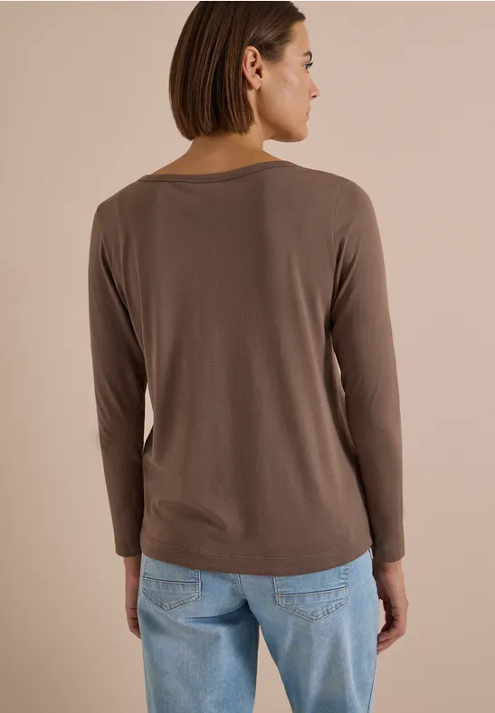 Frau von hinten in braunem Langarmshirt und blauen Jeans vor neutralem Hintergrund.