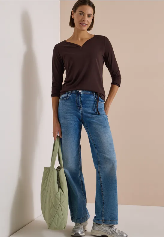 Frau in braunem V-Shirt, weiten Jeans und weißen Sneakers lehnt an hellem Hintergrund und hält eine grüne Tasche.