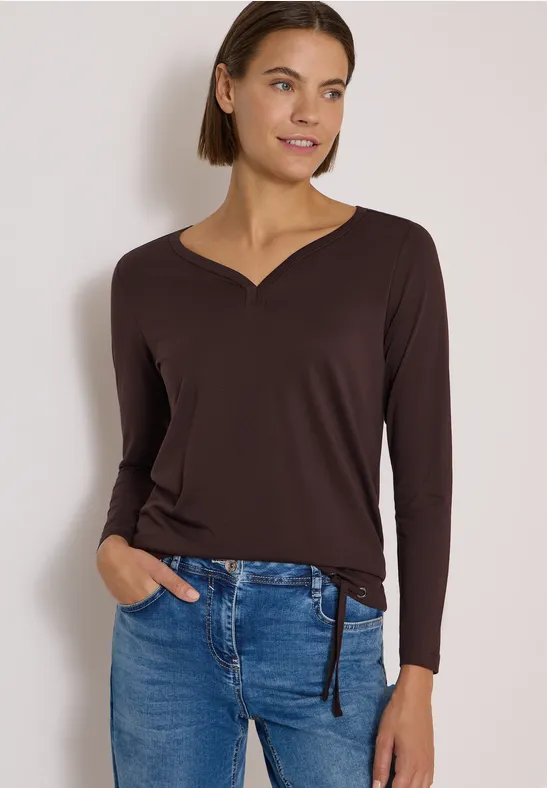 Frau trägt braunes Langarmshirt mit V-Ausschnitt und Jeans, eine Hand in der Tasche, lächelnd vor hellem Hintergrund.