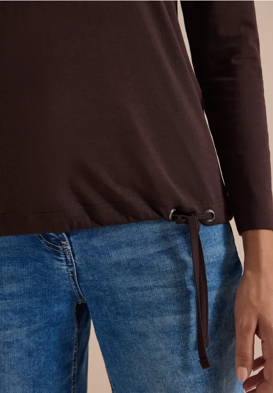 Teilsicht einer Person mit braunem Langarmshirt mit seitlichen Bändern und blauen Jeans.