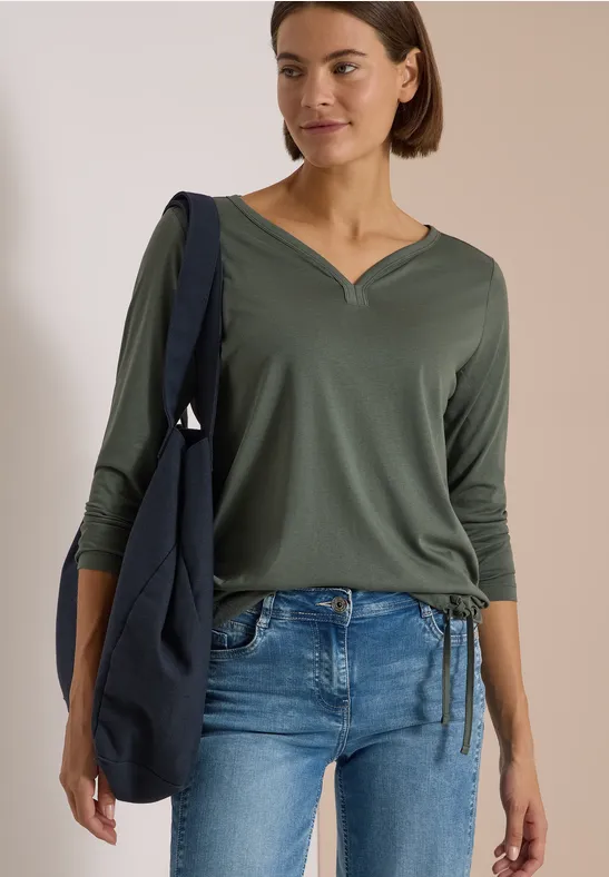 Frau in grüner V-Ausschnitt-Bluse mit 3/4-Ärmeln, blauer Jeans und dunkelblauer Umhängetasche.