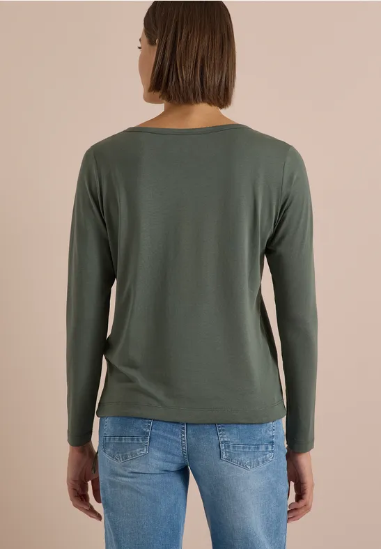 Person mit braunen Haaren von hinten, trägt grünes Langarmshirt und blaue Jeans vor beigem Hintergrund.