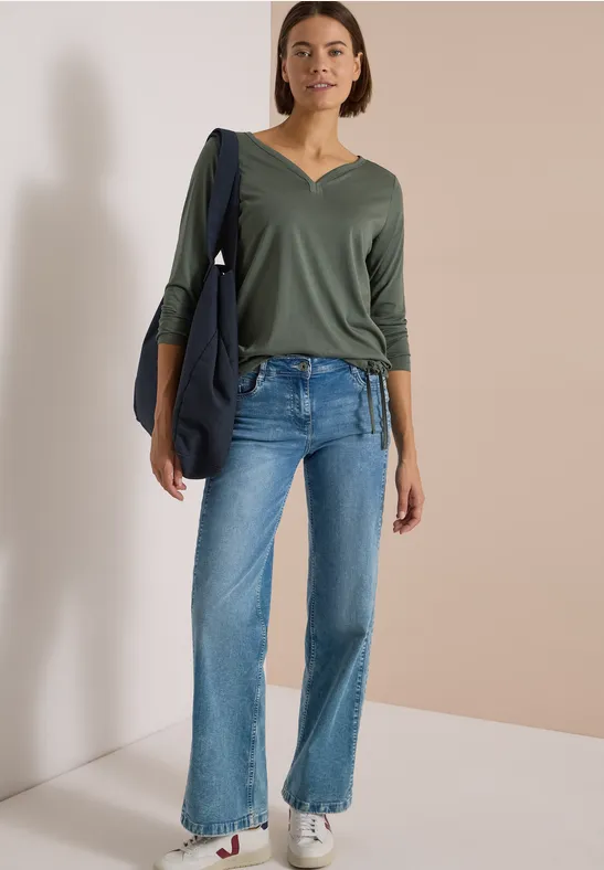 Eine Frau trägt grüne V-Neck-Bluse, blaue Jeans und weiße Sneaker, mit einer dunkelblauen Tasche über der Schulter.