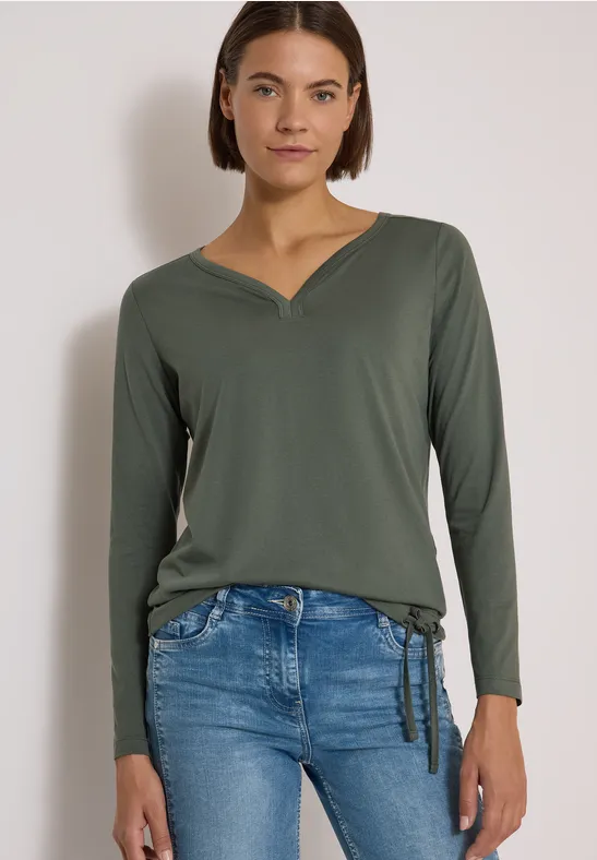 Frau trägt khakigrünes Langarm-Shirt mit V-Ausschnitt und helle Jeans mit seitlicher Schleife.