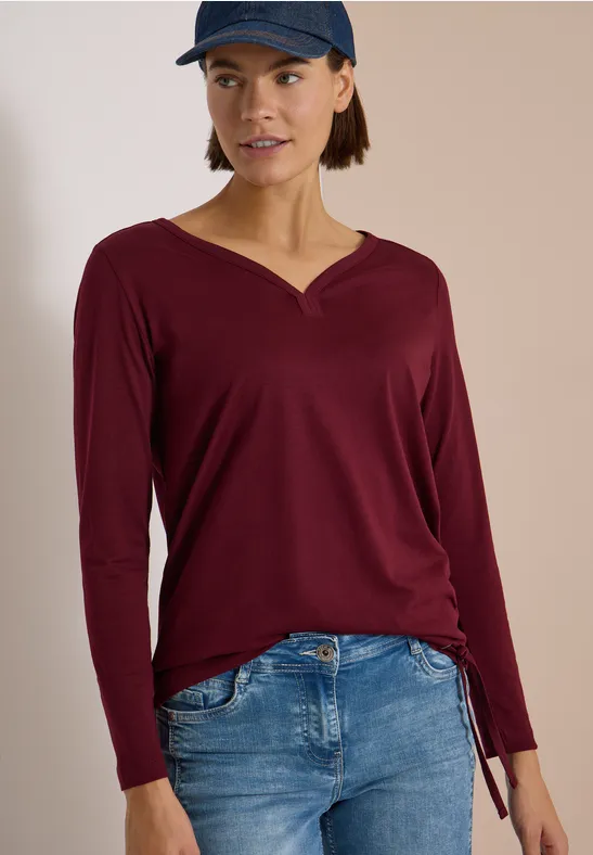 Frau trägt burgunderfarbenes Langarmshirt mit V-Ausschnitt und Tunnelzug, blaue Jeans und Cap.