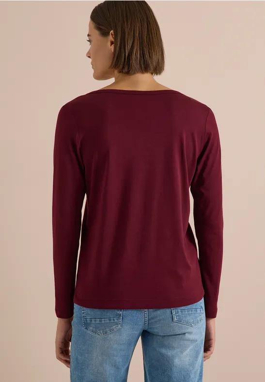 Frau von hinten mit langärmeligem bordeauxrotem Shirt und blauen Jeans vor neutralem Hintergrund.