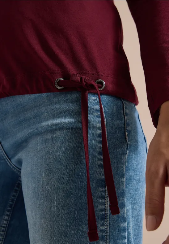 Person trägt blaue Jeans und ein dunkelrotes Langarmshirt mit Tunnelzug am Saum, Hand seitlich sichtbar.