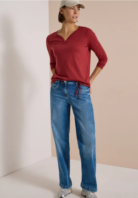 Frau im roten 3/4-Shirt, blauen Jeans und weißer Baseballkappe vor neutralem Hintergrund.
