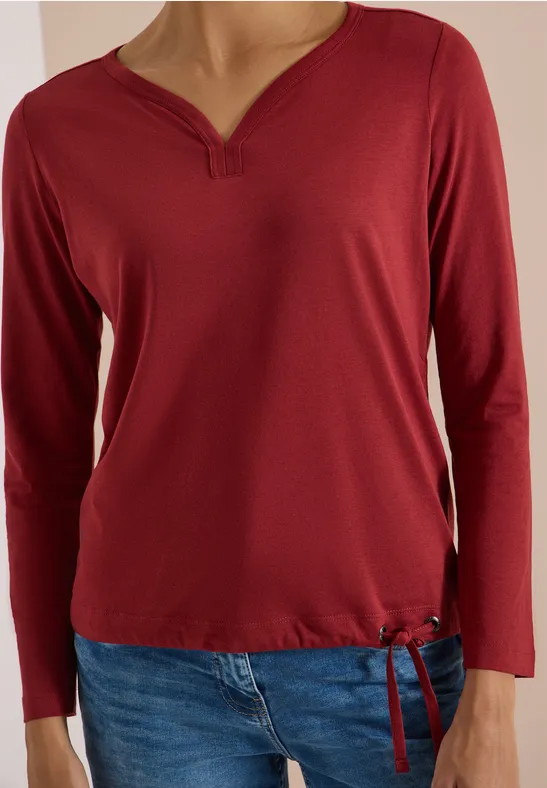Rotes Langarmshirt mit V-Ausschnitt und Kordelzug am Saum, dazu blaue Jeans.