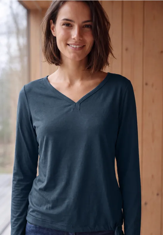 Junge Frau in blauem V-Ausschnitt-Langarmshirt lächelt vor Holzverkleidung mit Fensterblick.