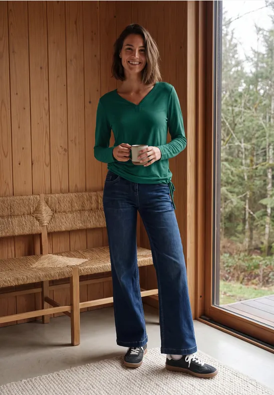 Een vrouw in een groen bloesje en spijkerbroek, glimlachend met een mok in een lichte houten ruimte bij een raam.