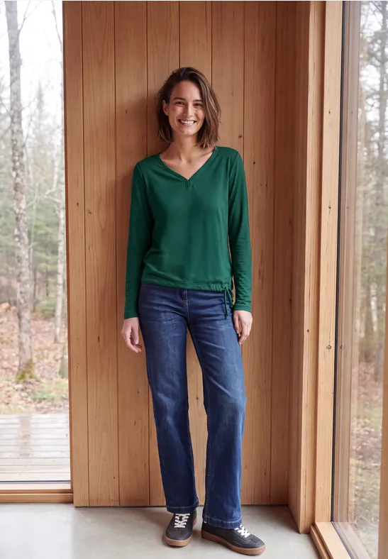 Een glimlachende vrouw in groen shirt en spijkerbroek staat voor een houten wand bij een raam.