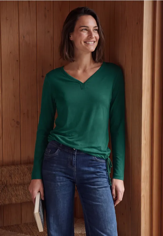 Een lachende vrouw in groene top en spijkerbroek staat in een houten ruimte en houdt een boek vast.