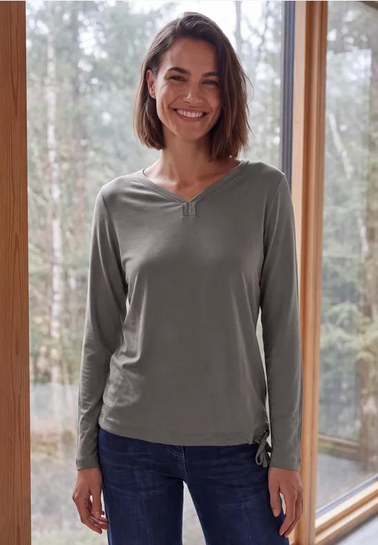 Lächelnde Frau mit schulterlangem Haar, grauem Langarmshirt und Jeans steht vor großen Fenstern mit Waldblick.