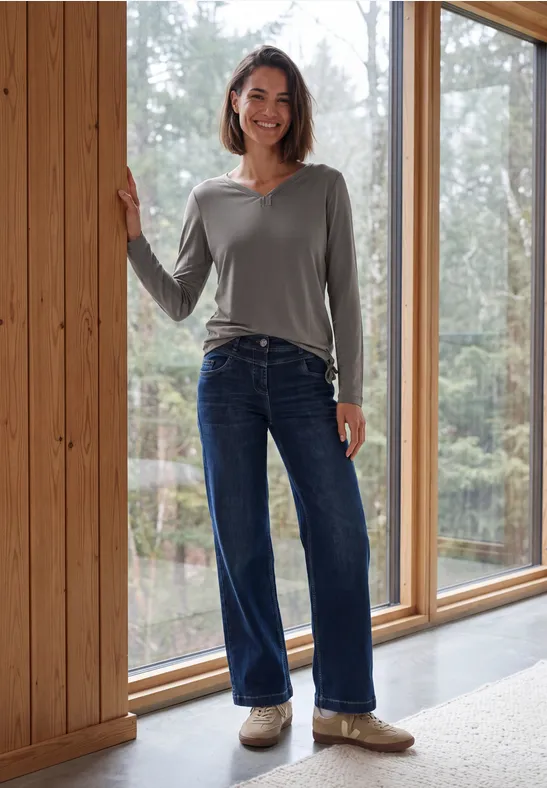 Frau in grauem Pullover und Jeans steht lächelnd vor großen Fenstern in einem Holzhaus im Wald.