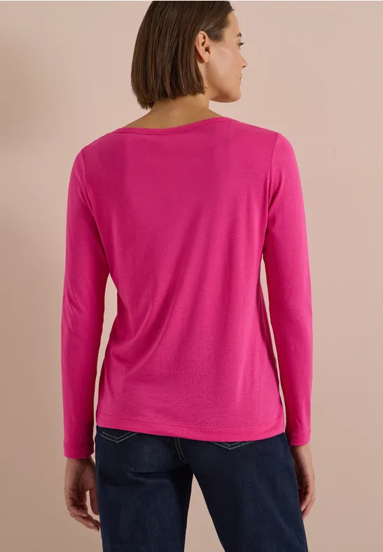Eine Frau von hinten in pinkem Langarmshirt und blauen Jeans vor neutralem Hintergrund.