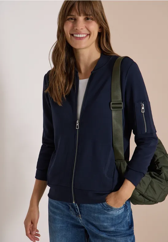Eine lächelnde Frau in blauer Strickjacke, Jeans und weißem Shirt, mit grünem Rucksack.