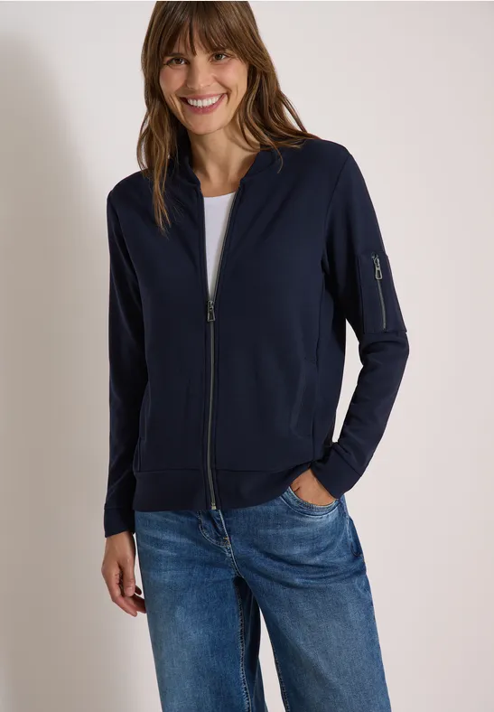 Lächelnde Frau in dunkelblauer Zip-Jacke und Jeans mit Händen in den Taschen vor weißem Hintergrund.
