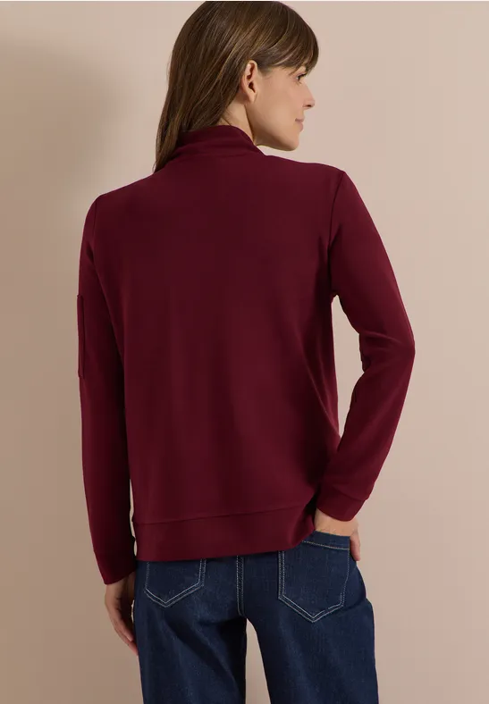 Rückenansicht einer Person in burgunderrotem Rollkragenpullover und Jeans vor beigem Hintergrund.