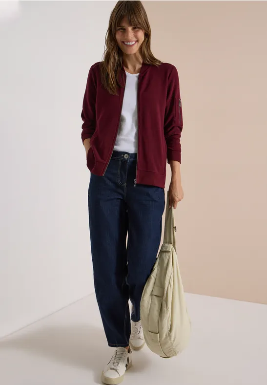 Frau trägt burgunderrote Jacke, weißes Shirt, lockere Jeans, weiße Sneaker und hält eine beige Tasche.