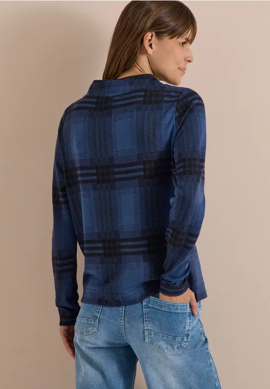 Frau von hinten in blau karierter Bluse und Jeans, Hände in den Hosentaschen.