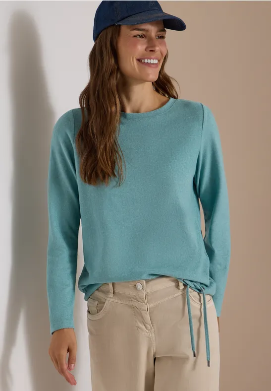 Frau im türkisfarbenen Pullover mit Seitenschnürung, beige Hose und Cap, lächelnd vor neutralem Hintergrund.