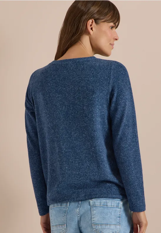Frau von hinten in dunkelblauem Strickpullover mit langen Ärmeln und Jeans vor schlichter Wand.