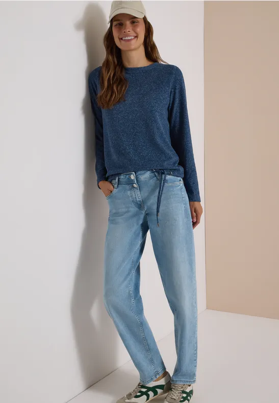 Junge Frau mit Baseballcap, blauem Pullover und Jeans lehnt lässig an der Wand und lächelt.