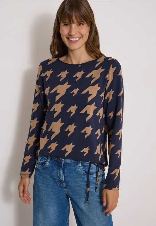 Eine lächelnde Frau trägt ein blau-beiges Hahnentritt-Longsleeve zu Jeans.