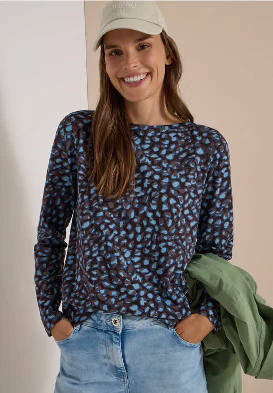 Une femme souriante portant un haut à manches longues imprimé, un jean, une casquette et une veste verte.