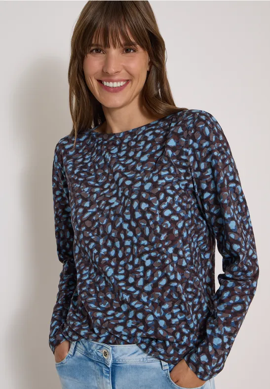 Une femme souriante en chemise marron à motifs bleus et jean clair.