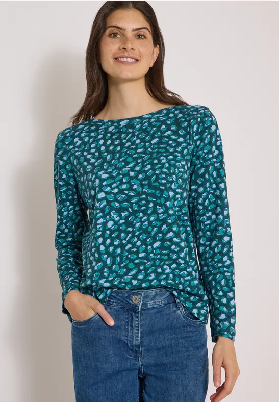 Femme souriante en t-shirt vert-bleu à motifs et jean bleu, mains dans les poches.