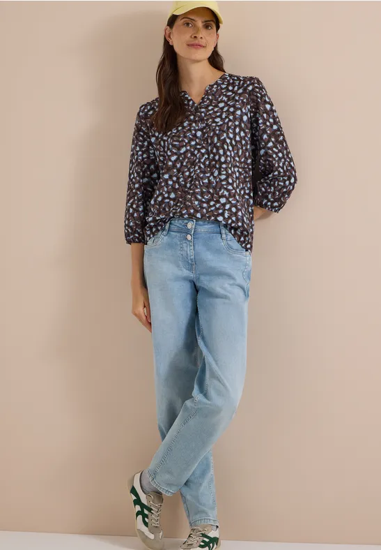 Femme en blouse à motifs, jean ample, baskets et casquette jaune devant fond beige