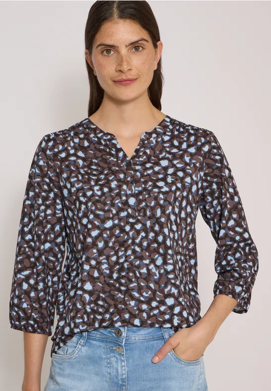 Femme portant une blouse à motif floral bleu et un jean, fond clair.
