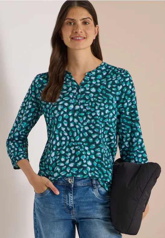 Femme souriante en chemisier vert à motifs et jean tenant une veste matelassée noire.