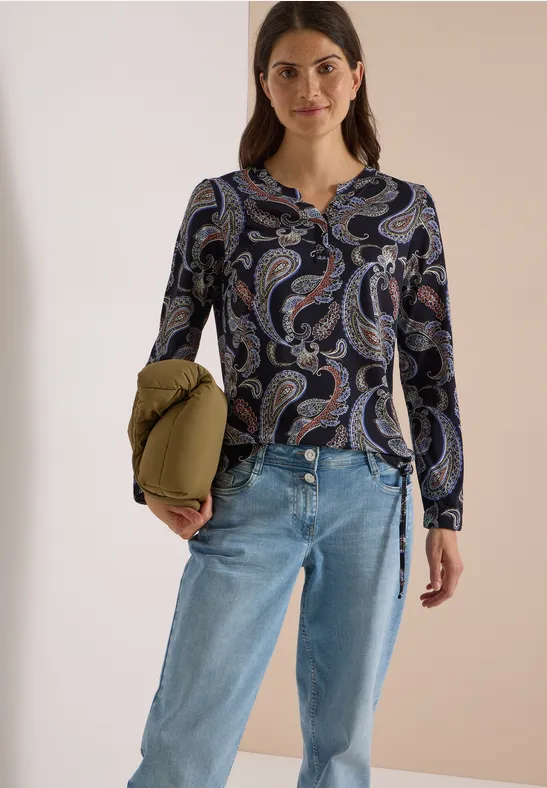 Eine Frau mit Brille trägt eine blaue Paisley-Bluse, helle Jeans und hält eine olivgrüne Jacke.