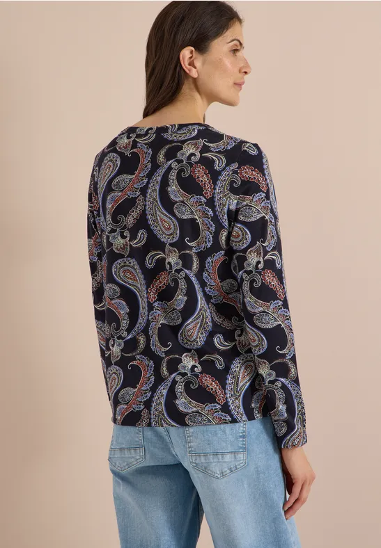 Frau von hinten in Jeans und dunkelblauem Paisley-Oberteil vor hellem Hintergrund.