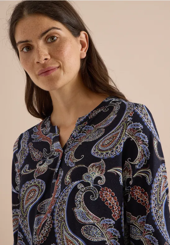 Frau mit dunklen Haaren in dunkelblauer Bluse mit buntem Paisley-Muster vor hellem Hintergrund.