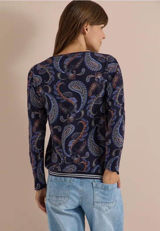 Rückenansicht einer Frau in dunkelblauem, paisley-bedrucktem Langarmshirt und hellblauen Jeans.
